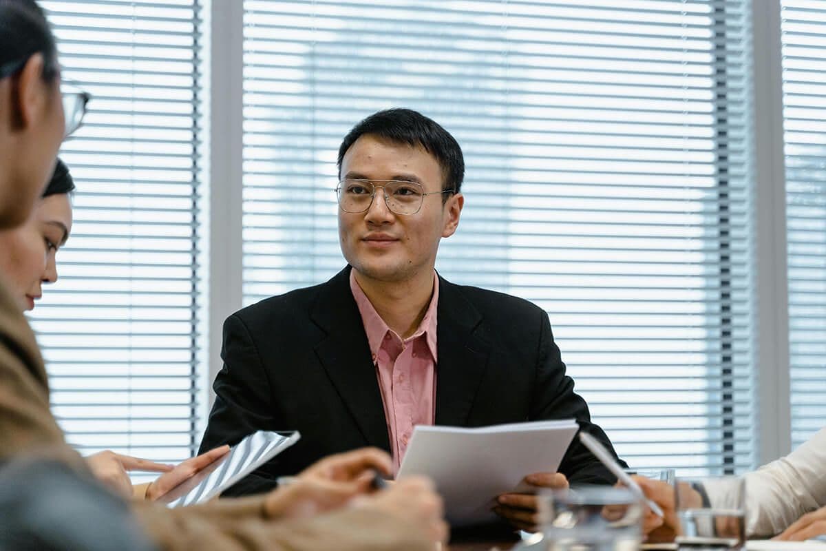 Man sitting at a table in a conference room in an office looking at colleagues as they explain a document he's holding.