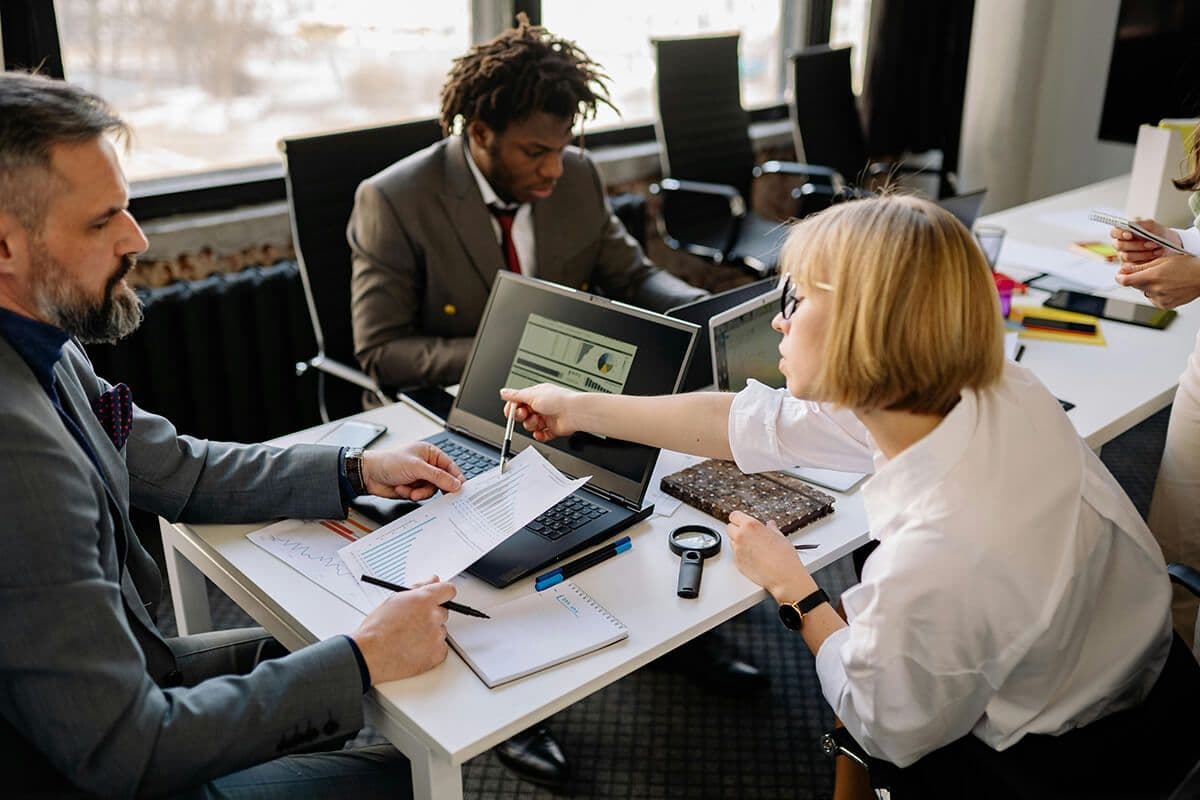 Woman showing male colleague data on a piece of paper. They're sitting at a long conference table with other employees.
